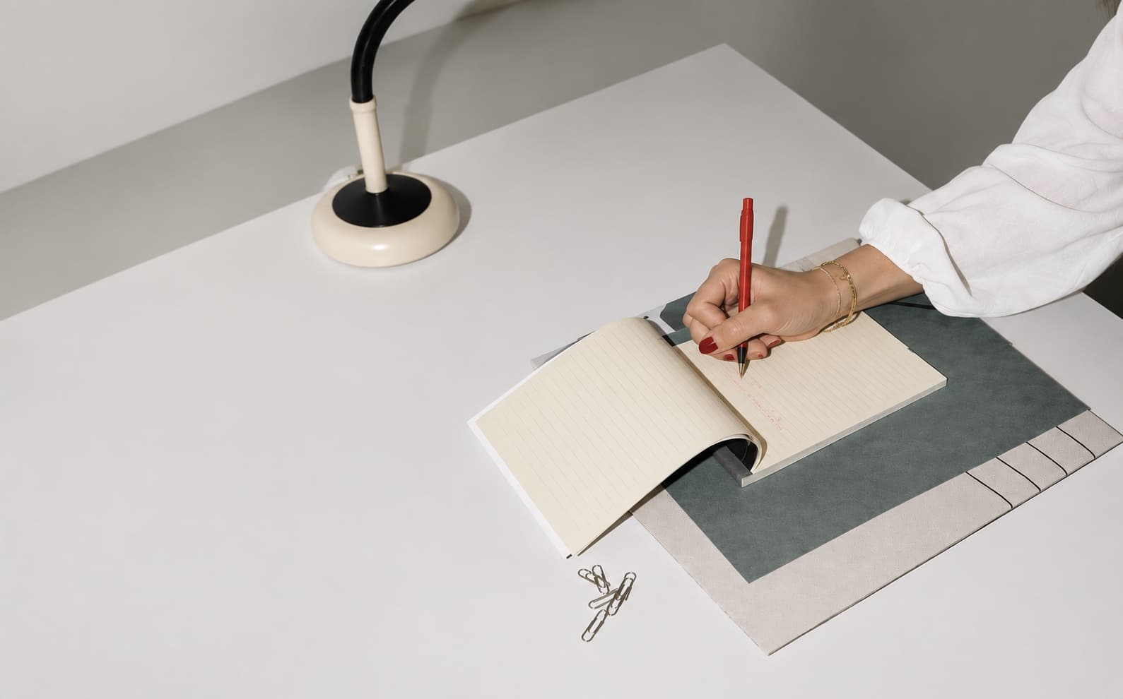 A hand writing in a notebook with a red pen beside a cream desk lamp