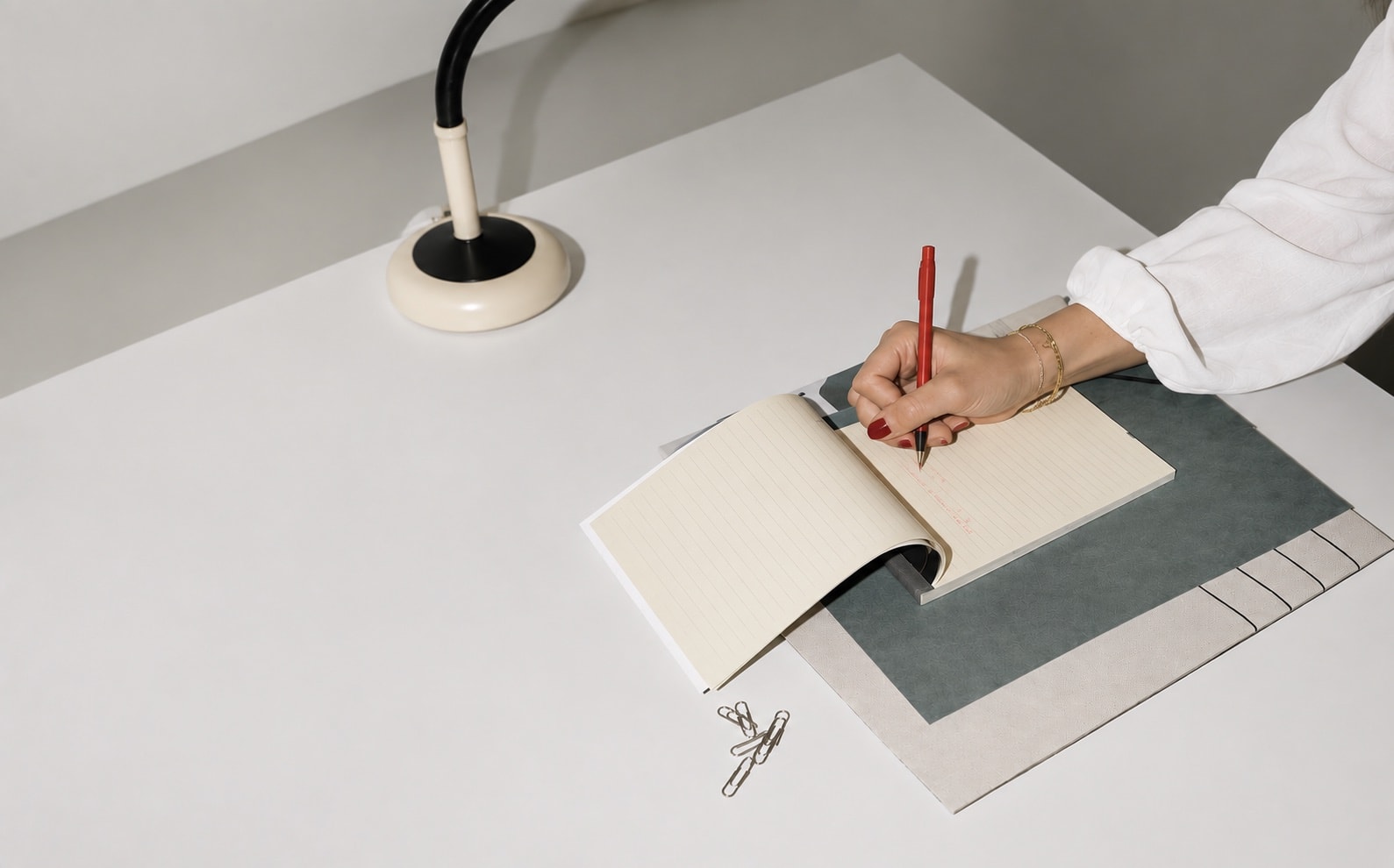 A hand writing in a notebook with a red pen beside a cream desk lamp